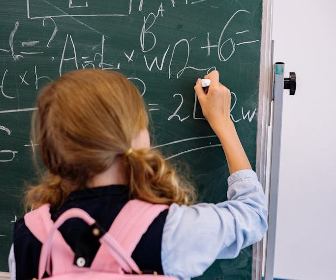 A young girl writes on a chalkboard, solving math equations in a classroom setting.