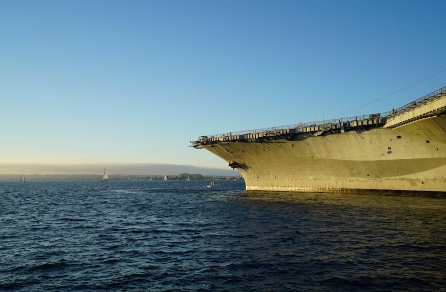 A large aircraft carrier docked in a serene harbor, surrounded by a clear blue sky and tranquil waters.