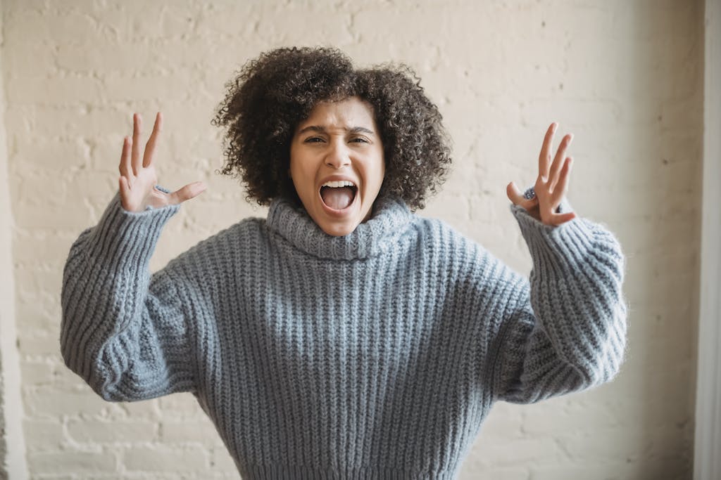 Irritated African American female with raised arms looking at camera while shouting loudly near wall in light room at home